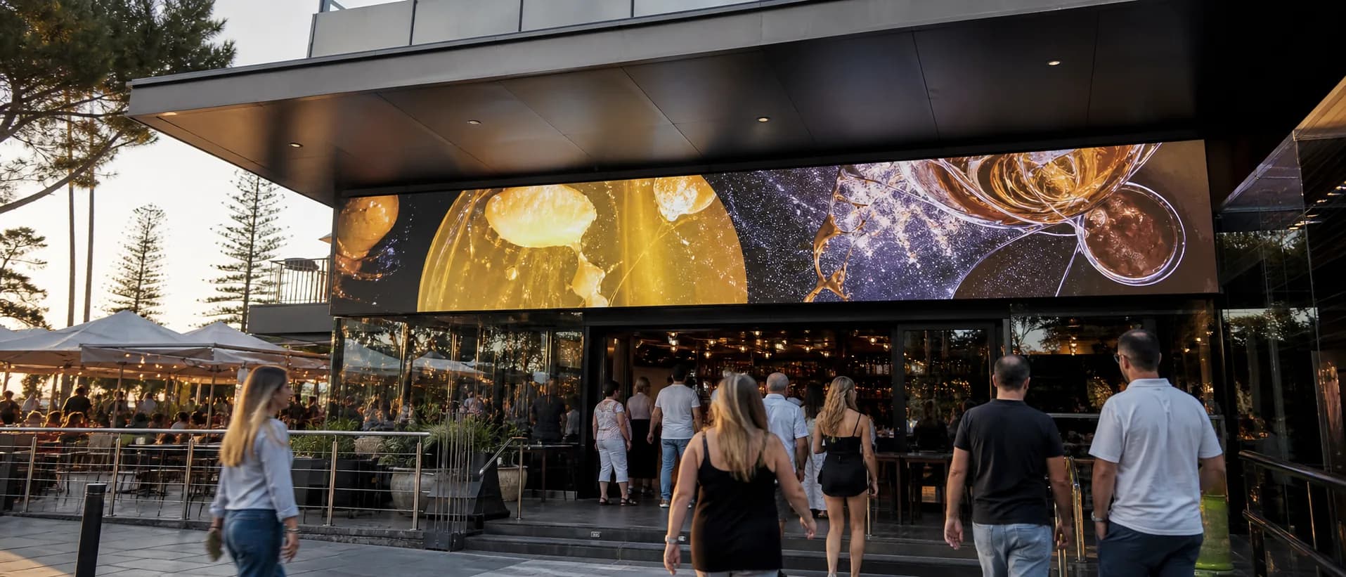Wide outdoor LED display above a hospitality venue entrance at dusk